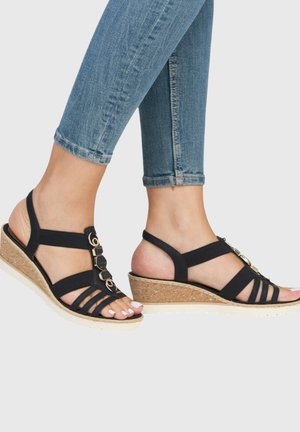 Pieds portant des sandales compensées noires à brides avec semelles en liège, associées à un jean skinny bleu retroussé.