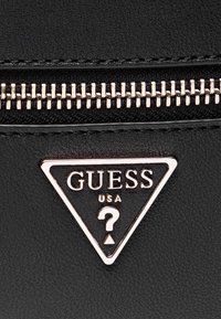 Surface en cuir noir avec fermeture éclair argentée et logo triangulaire Guess USA avec un point d'interrogation, détails de couture visibles.