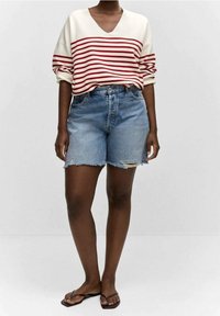 Henkilö, jolla on päällä punavalkorahallinen pitkähihainen paita, kuluneet siniset farkkushortsit ja mustat sandaalit, seisoo tasaisessa taustassa.