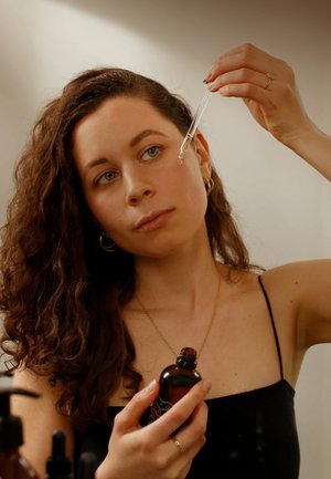 Ein Glas-Pipette wird über ein Gesicht gehalten und gibt eine klare Flüssigkeit ab. Die Person hält eine dunkelbernsteinfarbene Glasflasche mit dekorativen Akzenten. Schwarze Kleidung.