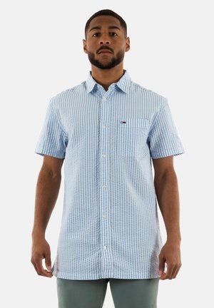 Camisa de manga corta a rayas en azul claro y blanco, con un bolsillo en el pecho frontal y un pequeño parche de logo en el lado izquierdo.