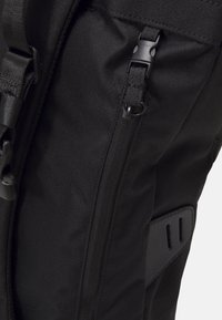 Musta reppu, jossa on teksturoitu ja kestävä kangas; siinä on vetoketjullinen suljin ja säädettävät hihnat; mukana on sivutasku, jossa on sileä paneeli.