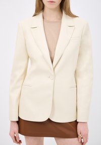 Blazer su misura color crema con rever a punta, chiusura a un bottone e due tasche laterali. Texture liscia, design strutturato.