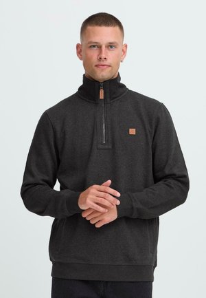 Man with short hair wearing a dark gray quarter-zip pullover sweatshirt, standing with hands clasped in front against a plain background.
