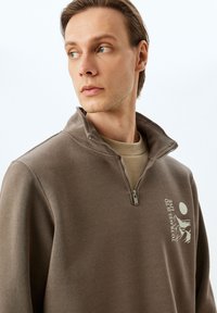 Brauner Zip-Hoodie mit hohem Kragen, ausgestattet mit einem Front-Design und strukturiertem Stoff. Darüber getragen wird ein beiges Rundhals-Shirt.