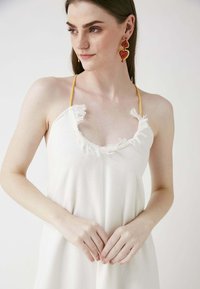 Weißes Kleid mit Fransenverzierungen am Ausschnitt, gelben Trägern und einer lockeren Passform. Das Model trägt große rote und goldene Ohrringe.