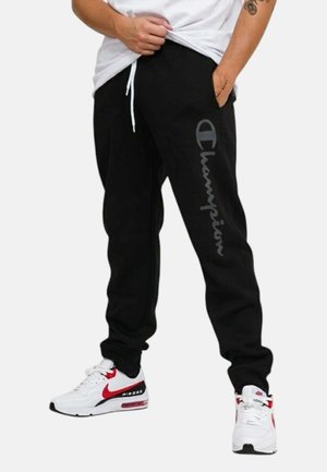 Persona che indossa pantaloni della tuta Champion neri con logo laterale e sneakers Nike Air Max bianche con accenti rossi, in piedi con le mani in tasca.