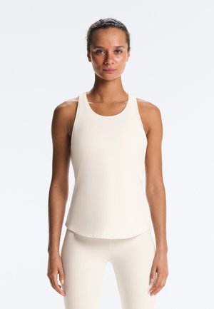 Kvinde med opsat hår iført en ærmeløs, lysebeige atletisk top og matchende leggings, stående mod en ensfarvet hvid baggrund.