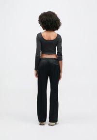 Ženska s kodrastimi lasmi, oblečena v črn kratek pulover z dolgimi rokavi, črne hlače in bele superge, stoji s pogledom stran na belem ozadju.