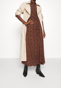 Beige trenčkot přes černé květinové maxi šaty s červenými a růžovými květy. Model má na sobě černé špičaté kotníkové boty.