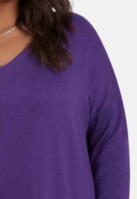 Lila stickad tröja med V-ringning och långa ärmar. Mjuk textur och ribbad detalj synlig på tyget.