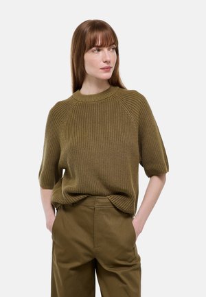 Jeune femme aux cheveux bruns et lisses portant un pull en maille vert olive à manches courtes et un pantalon taille haute assorti, mains dans les poches.