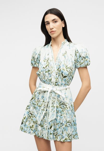 Woman with long dark hair wears a short white dress with blue floral print, puff sleeves, and a tied waist belt, standing against a plain background.