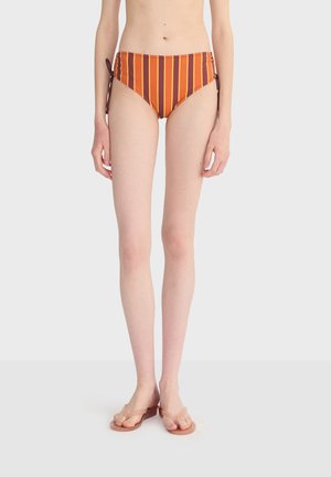 Žena u visokim badićima sa vertikalnim prugama u narančastoj, crvenoj i žutoj boji, stoji bosa na bijeloj pozadini.