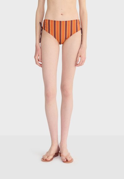 Femme portant un bas de bikini taille haute avec des rayures verticales orange, rouge et jaune, debout pieds nus sur un fond blanc.
