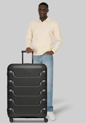 Mann in beige langärmeligem Hemd und Jeans steht und hält den Griff eines großen schwarzen, strukturierten Hartschalenkoffers auf Rollen.