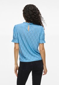 Lyseblå bluse med pufferm, perforert mønster og en nøkkelhullsåpning bak. Laget av et strukturert stoff.