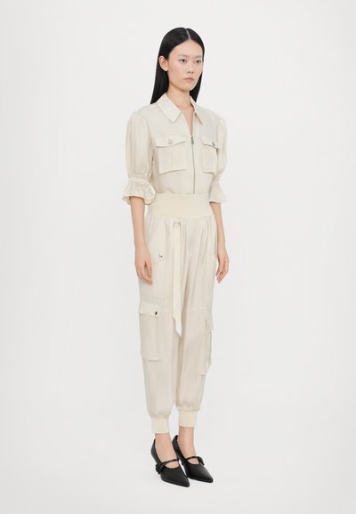 Cremefarbener Jumpsuit mit Reißverschluss vorne, puffigen Ärmeln, zwei Brusttaschen und schmalen Bündchen. Kombiniert mit schwarzen spitzen Absatzschuhen.