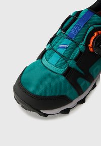 Sportssko med overdel i teal mesh, svarte detaljer, teksturert såle og en sirkulær justeringsknapp. Har kontrasterende kanting og søm.