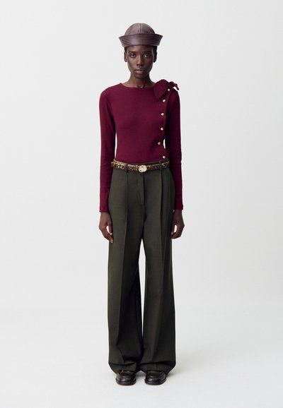 Top lungo color bordeaux con dettagli di bottoni e un'accattivante fiocchetto, abbinato a pantaloni larghi a vita alta color verde oliva e un cappello marrone testurizzato.