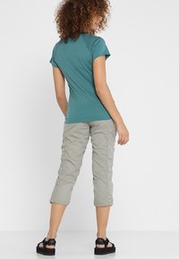 Kobieta z kręconymi włosami, ubrana w turkusową koszulkę z krótkim rękawem, jasnoszare spodnie 3/4 i czarne sandały na platformie, stojąca na tle białym.