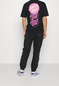 Svart t-shirt med stor rosa grafisk text "SANTA CRUZ" tryckt på ryggen. Lös passform, kombinerad med mörka byxor och flerfärgade sneakers.