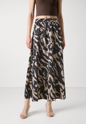 Vrouw draagt een lange beige en zwart abstract patroon rok, een kort bruin topje en doorzichtige sandalen met hoge hakken, staand tegen een effen achtergrond.