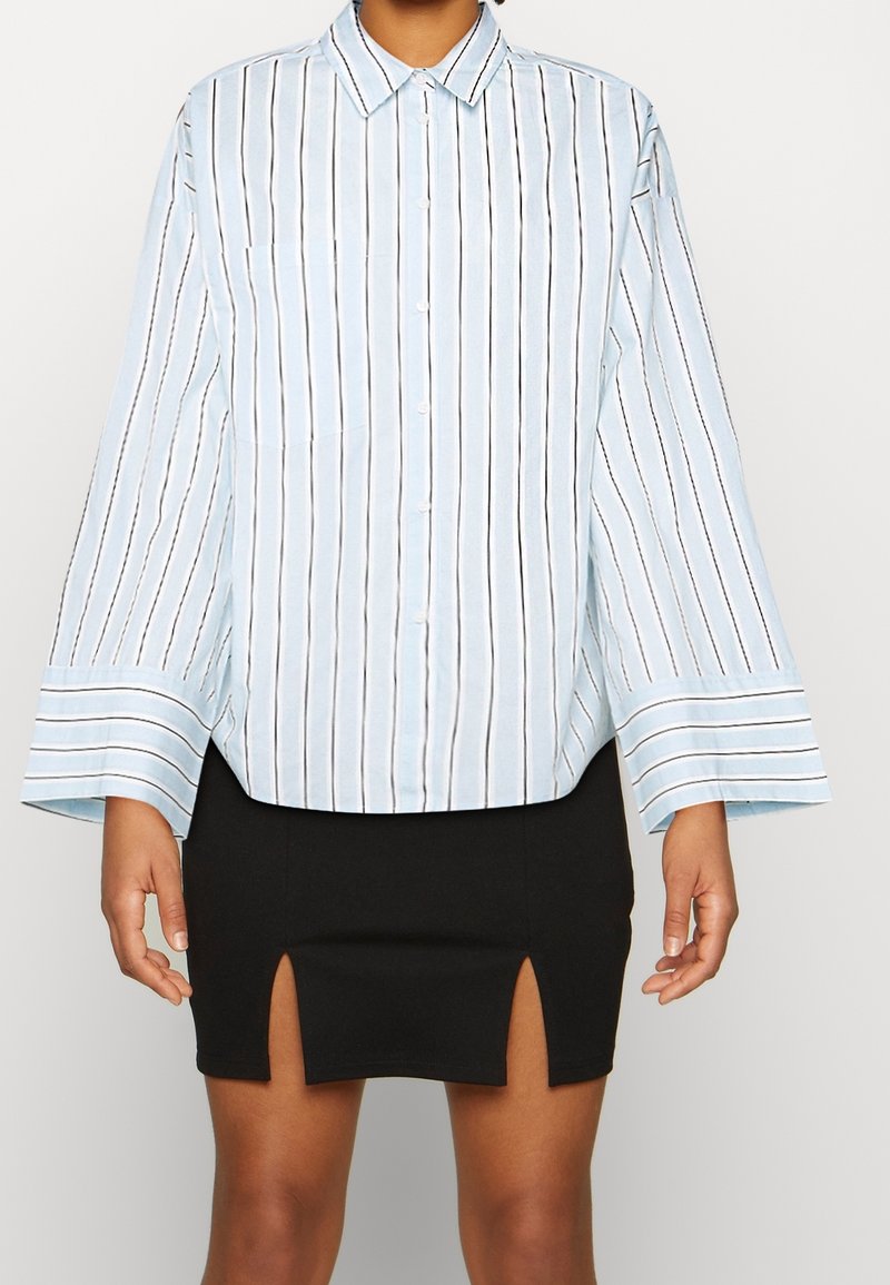 Light blue button-up shirt with vertical black stripes, wide sleeves, and a chest pocket, paired with a black mini skirt with side slits.