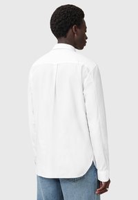 Camicia bianca a maniche lunghe con piega sulla schiena e colletto a punta, abbinata a jeans in denim azzurro, vista da dietro.
