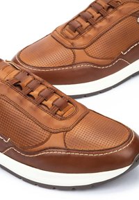 Sneaker in pelle marrone con texture perforata, suola intermedia bianca a contrasto e lacci marroni. Presenta una punta arrotondata e dettagli di cucitura sottili.