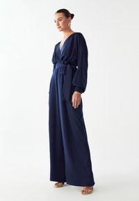 Marineblauwe jumpsuit met een diepe V-hals, lange mouwen en een gestrikte taille. Gladde stof met wijde, vloeiende pijpen. Gouden accent sandalen.