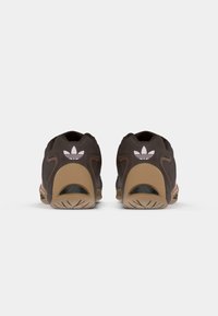 Tmavohnedé športové topánky s textúrovanou tkaninou, gumovou podrážkou a ružovým logom Adidas na päte. Zaoblený dizajn chrbta s výrezmi.