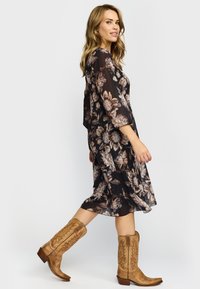 Kvinne går iført en svart blomstrete midi-kjole med gjennomsiktige ermer og brune broderte cowboystøvler mot en hvit bakgrunn.