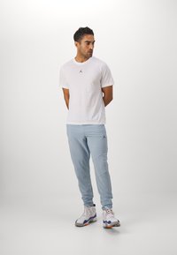 Hvid kortærmet skjorte med sort logo, lyseblå bukser med logo på benet, og hvide sneakers med blå og orange accenter.