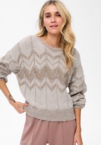 Vrouw met lang blond haar, gekleed in een beige gebreide trui met patroon en mauve broek, poseert met één hand in de zak tegen een witte achtergrond.