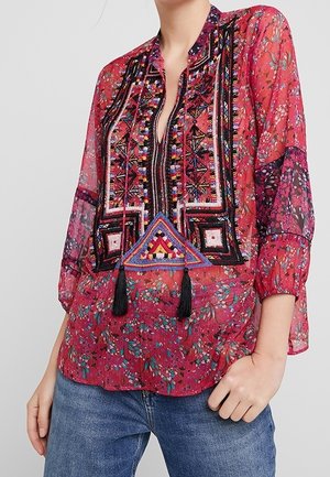 Červená průsvitná halenka s květinovými vzory, detailním vyšíváním a rozparkem na výstřihu. Má třásně a volné rukávy. Kombinováno s modrými džíny.