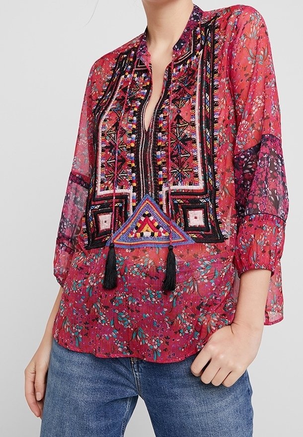 Blouse rouge transparent avec motifs floraux, broderies détaillées et encolure fendue. Dotée de pompons et de manches bouffantes. Associée à un jean bleu.