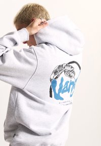 Felpa con cappuccio grigio chiaro realizzata in materiale morbido, con una grafica nera e blu sul retro che raffigura una mano e un testo di accento.