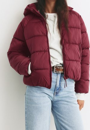 Personne portant une doudoune bordeaux par-dessus une chemise blanche à manches longues et un jean bleu clair avec une ceinture marron cloutée.