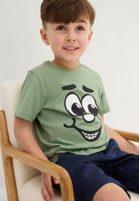 T-shirt verde in cotone con una grande grafica di un volto di cartone animato nero sul davanti. Maniche corte e scollo rotondo.