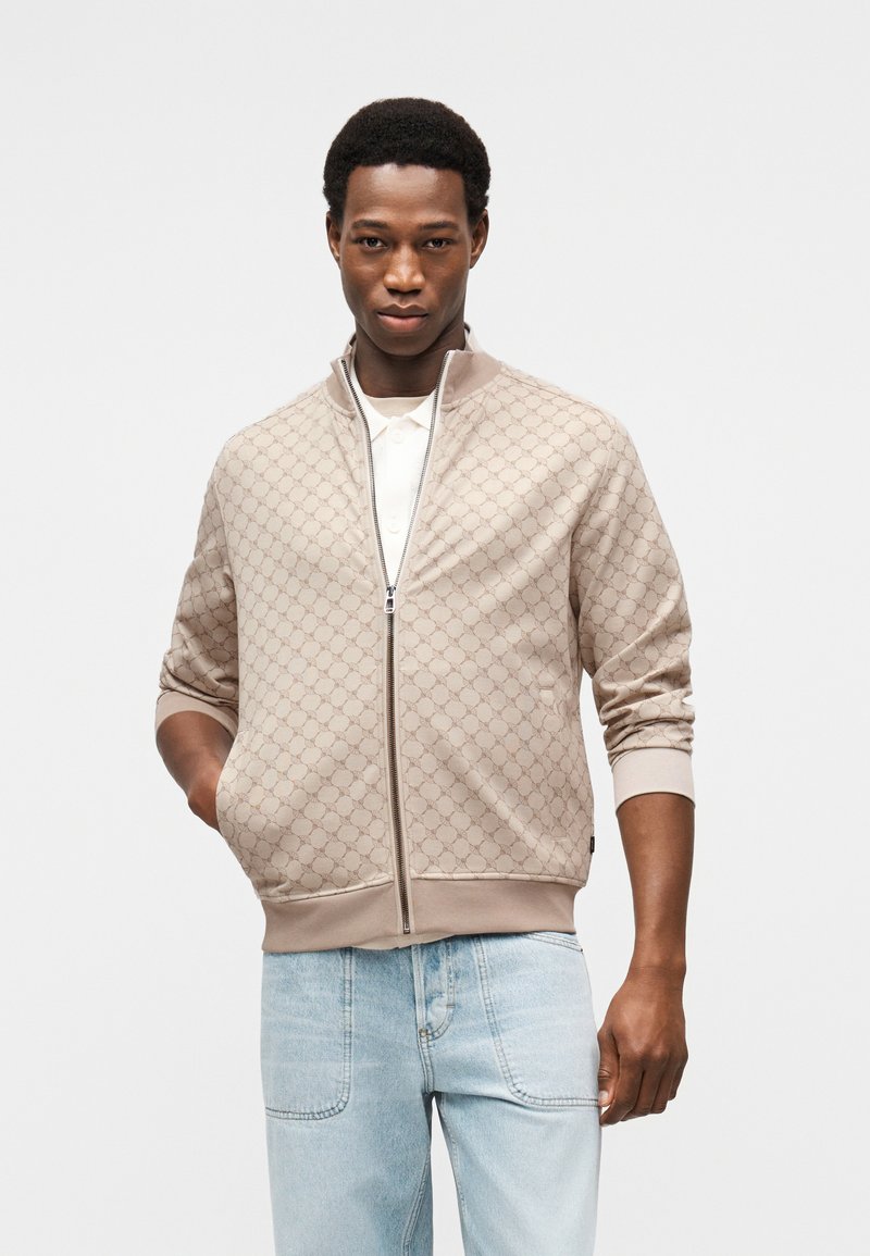 Veste beige zippée avec un motif embossé intégral, poignets et ourlet côtelés, fabriquée en tissu doux, portée avec un jean bleu clair.