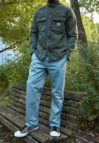 Oliwkowa koszula z długim rękawem i dwoma kieszeniami na piersi; jasnoniebieskie spodnie; czarno-białe trampki; na zewnątrz, siedzący na drewniej ławce.