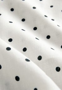 Weißer Stoff mit strukturierter Oberfläche, der ein Muster gleichmäßig verteilter schwarzer Punkte unterschiedlicher Größe zeigt. Weiches und leichtes Material.