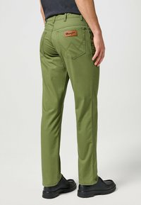 Groene katoenen broek met een rechte pijp, zichtbare stiksels en een leren patch met "Wrangler" op de achterzak. Zwarte schoenen eronder gestyled.
