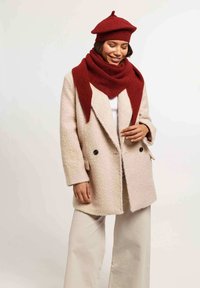 Abrigo beige texturizado con botones negros, combinado con una bufanda roja gruesa y un boina a juego. Modelo vistiendo pantalones de pierna ancha en color crema.