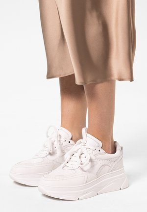 Jambes portant une jupe beige et des baskets blanches épaisses avec des détails en texture tissée, debout sur un fond blanc uni.