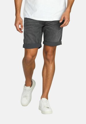 Mann mit hochgekrempelten dunkelgrauen Jeansshorts, weißen Sneakern und weißem Hemd, der vor einem schlichten hellen Hintergrund läuft.