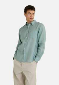 Chemise à manches longues vert clair en tissu texturé, avec un devant à boutons et une poche poitrine ; associée à un pantalon beige.