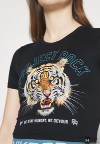 Svart T-shirt med en rytande tigergrafik, blå text "PROJECT ROCK" ovanför och vit text "WE STAY HUNGRY, WE DEVOUR" under tigerns ansikte.