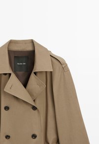 Beiger Trenchcoat aus Baumwollmischung, mit doppelreihigen Knöpfen, weitem Kragen und Schulterklappen. Etikett innen sichtbar.